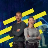 Eine Ermittlerin und ein Ermittler der Kriminalpolizei sind im Vordergrund zu sehen. Sie tragen zivile Kleidung. Hinter ihnen ist eine Grafik mit gelben Streifen zu sehen. Im Hintergrund ist abgedunkelt und verschwommen ein Polizeifahrzeug auf einer Straße zu sehen.