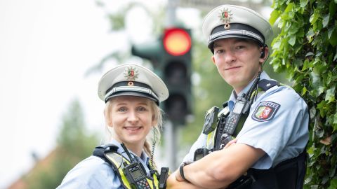 Uniformierte Polizistin links, uniformierter Polizist rechts, beide tragen eine weiße Polizeimütze auf dem Kopf, eine blaue Schutzweste, Funkgerät, Waffe im Holster und Pfefferspray. Im Hintergrund ist verschwommen eine rot zeigende Ampel und ein Stoppschild zu sehen.